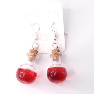 May include: A pair of dangle earrings featuring small glass bottles with cork stoppers filled with a red liquid. The bottles are attached to silver hooks.