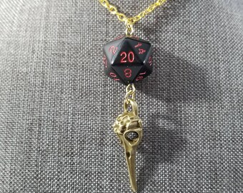 May include: A gold-colored chain necklace featuring a black D20 die pendant with red numbering and a gold-colored bird skull charm. The necklace is displayed against a gray background.