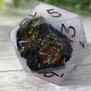Puede incluir: Un dado transparente de múltiples caras con números negros. El dado está relleno de un material brillante y colorido que refleja la luz. Es un artículo único y decorativo, perfecto para juegos de mesa o como objeto de colección.