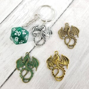 May include: A green 20-sided die with white numbers hangs from a silver key chain with a silver dragon charm. Three additional dragon charms are shown in silver, gold, and bronze.