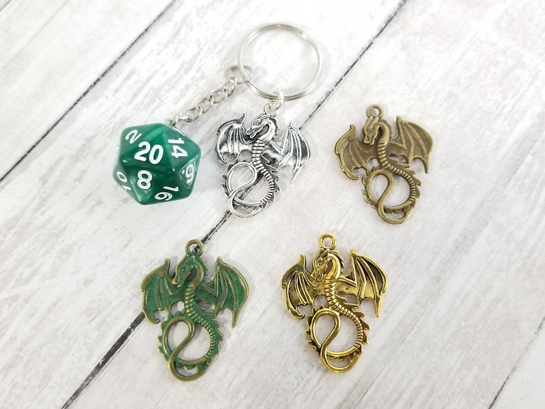May include: A green 20-sided die with white numbers hangs from a silver key chain with a silver dragon charm. Three additional dragon charms are shown in silver, gold, and bronze.