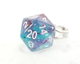 May include: A D20 die ring with a silver band. The die is a mix of teal, purple, and white, with the number 20 prominently displayed. The ring is designed for tabletop gaming enthusiasts.