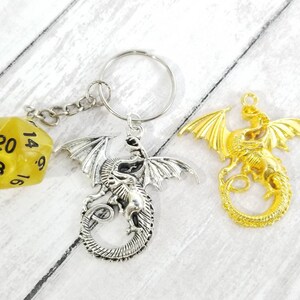 May include: A silver dragon keychain with a yellow 20-sided die attached. The dragon is curled up with its wings spread out. There is also a gold dragon keychain with its wings spread out.