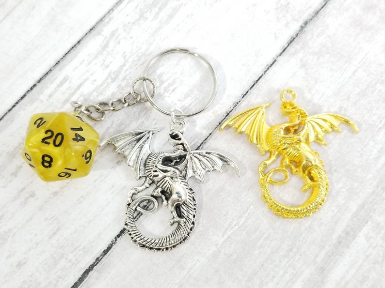 May include: A silver dragon keychain with a yellow 20-sided die attached. The dragon is curled up with its wings spread out. There is also a gold dragon keychain with its wings spread out.