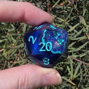 May include: A twenty-sided die with a dark blue, glittery interior and teal numbering. The die is held between two fingers against a backdrop of green foliage. The number 20 is prominently displayed.