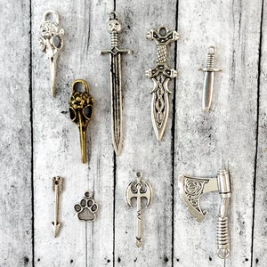 May include: A collection of silver and bronze charms featuring a variety of designs, including swords, axes, arrows, and animal paw prints.