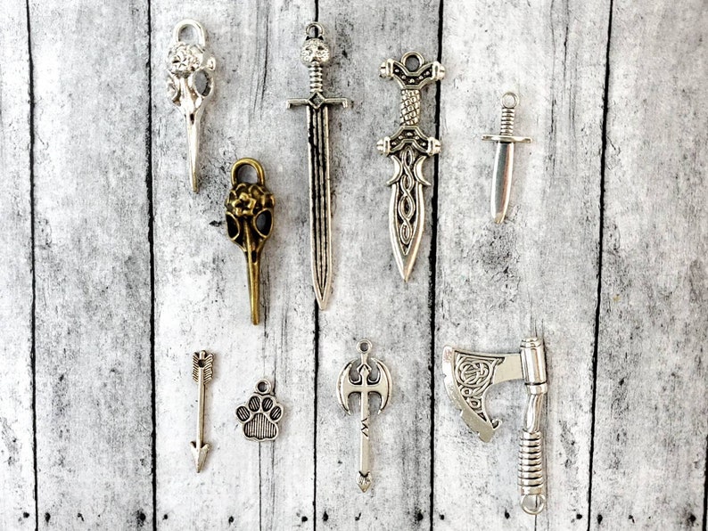 May include: A collection of silver and bronze charms featuring a variety of designs, including swords, axes, arrows, and animal paw prints.