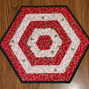 May include: A hexagonal red and white table runner with a candy cane pattern. The runner has a green border and is made of fabric.