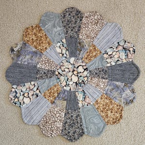 May include: A circular fabric mat featuring a patchwork design of various stone, wood grain, and marble patterns in shades of gray, brown, and tan. The mat has a scalloped edge and a central circular panel, all constructed from quilted fabric pieces.