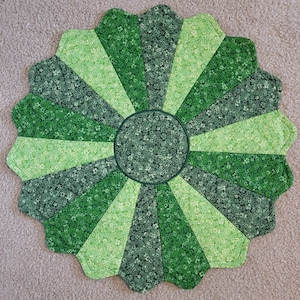 May include: A round, green quilted placemat with a scalloped edge. The design features a central circle and radiating wedge shapes in various shades of green, with floral patterns. The placemat is approximately 15 inches in diameter.