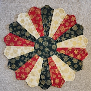 May include: A circular quilted fabric mat featuring a radial pattern of alternating wedge-shaped panels in black, red, and cream. Each panel is decorated with gold-colored snowflake motifs, and a central circular patch features matching snowflakes.