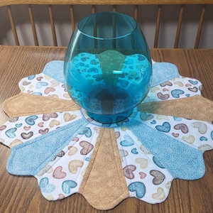 May include: A large, clear blue glass bowl sits on a decorative, flower-shaped placemat. The placemat features a pattern of hearts in shades of blue, brown, and white, with sections of solid blue and tan fabric. The bowl is empty.