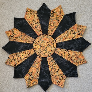 May include: A handmade, star-shaped quilted trivet. The trivet features a central circle and radiating points, with alternating black and paisley-patterned segments in shades of gold and orange. The paisley design includes swirls and dots.