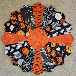 May include: A circular quilted fabric mat featuring a central orange marbled circle surrounded by twelve wedge-shaped panels. The panels display Halloween-themed patterns including orange pumpkins, black bats, white spiderwebs, skeletons, and white ghosts on black backgrounds.
