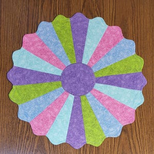 May include: A colorful, circular placemat with a scalloped edge. The design features alternating segments of pink, light blue, green, and purple radiating from a central purple circle. The placemat is likely made of fabric.