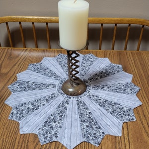 May include: A white candle sits on a metal candle holder with a spiral design. The candle holder is on a gray and white round table runner with a scalloped edge.