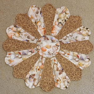 May include: A round, brown and white quilted table runner with a seashell design. The center of the runner features a circle of various seashells.