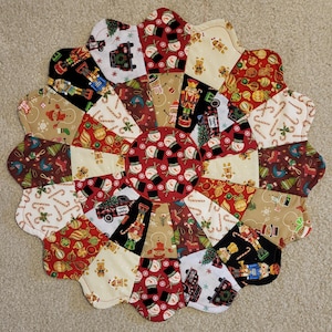 May include: A circular, quilted Christmas placemat with a scalloped edge. The patchwork design features festive patterns including snowmen, candy canes, nutcrackers, and Christmas trees in red, white, black, and tan. The center is a red circle with a snowman pattern.