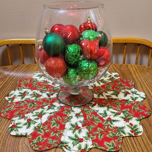 May include: A glass bowl filled with red and green Christmas ornaments sits on a white and red holiday table runner with a holly pattern.