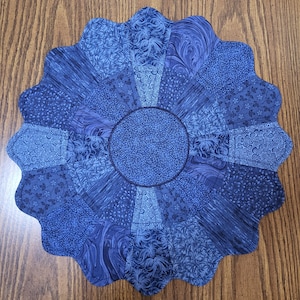 May include: A round, blue quilted placemat with a scalloped edge. The placemat features a central circle and radiating segments, each with a different blue patterned fabric. The fabrics include floral, swirl, and geometric designs, creating a visually interesting piece.