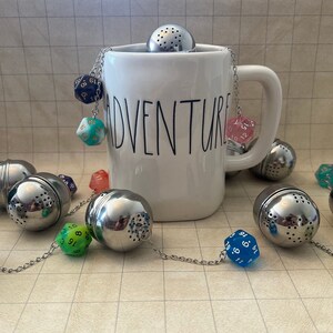 May include: A white mug with the word "Adventure" printed on it, with a chain of metal tea infusers and colorful dice attached. The mug is filled with tea infusers and dice.