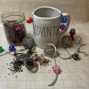May include: A white ceramic mug with the word "ADVENTURE" printed on it, surrounded by dice and tea infusers. A glass jar filled with dried rose petals is also present. The scene is set on a grid-patterned surface.