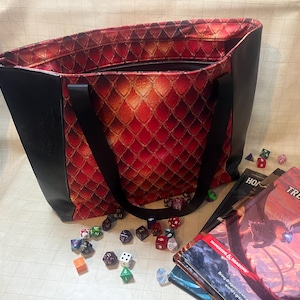May include: A large tote bag with a red dragon scale pattern interior and black exterior. The bag has black handles and a zipper closure. Several dice and Dungeons & Dragons books are next to the bag.