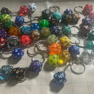 May include: A collection of colorful, multi-sided dice keychains. The dice are made of clear plastic with white numbers and gold accents. The keychains are attached to silver metal rings.