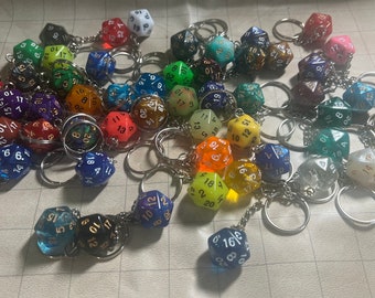 May include: A collection of colorful, multi-sided dice keychains. The dice are made of clear plastic with white numbers and gold accents. The keychains are attached to silver metal rings.
