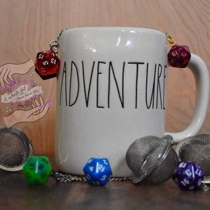 May include: A white ceramic mug with the word "Adventure" printed on it. The mug has a gold chain with a red, purple, and blue dice charm hanging from it. There are also three tea infusers with dice charms on a wooden surface.