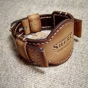 May include: A brown leather watch cuff with white stitching and a metal buckle. The cuff has the embossed text "SHERP" and "IGOR SHARIN". The watch cuff is designed to protect the watch face. The cuff is made of leather.