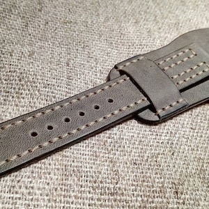 May include: A grey leather watch strap featuring a wide cuff design with light-colored contrast stitching. The strap includes a pointed end with five punched holes for adjustment and a matching leather keeper loop to secure the excess band length.