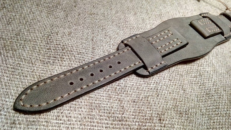 May include: A grey leather watch strap featuring a wide cuff design with light-colored contrast stitching. The strap includes a pointed end with five punched holes for adjustment and a matching leather keeper loop to secure the excess band length.