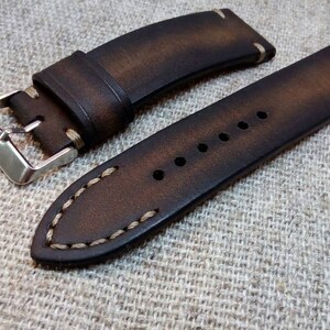 May include: A brown leather watch strap with white stitching. The strap has a silver buckle and is shown on a textured fabric background.