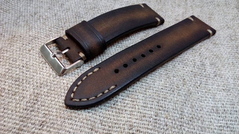 May include: A brown leather watch strap with white stitching. The strap has a silver buckle and is shown on a textured fabric background.