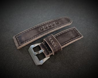 May include: A close-up of a brown leather watch strap with a silver buckle. The strap has a distressed appearance with visible stitching and the Roman numeral "XXXX" stitched on the strap. The watch band is shown on a dark gray surface.
