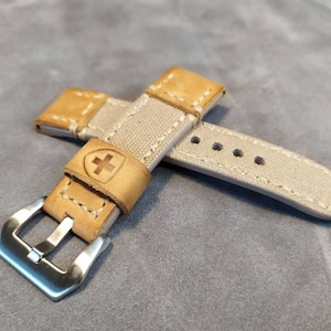 May include: A beige canvas watch strap with a brown leather buckle and a silver buckle clasp. The leather buckle has a small cross embossed on it.