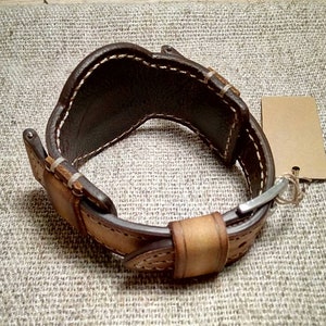 May include: A brown and tan leather watch cuff with white stitching and a metal clasp. The cuff has a wide design and a blank tag attached. The leather is hand-stitched.