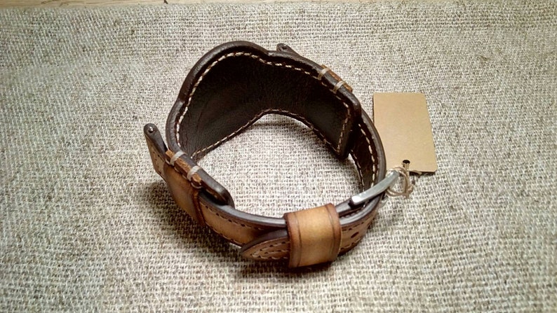 May include: A brown and tan leather watch cuff with white stitching and a metal clasp. The cuff has a wide design and a blank tag attached. The leather is hand-stitched.