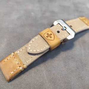 May include: A beige canvas watch strap with a brown leather trim and a silver buckle. The strap has a stitched design and a small shield with a cross logo.