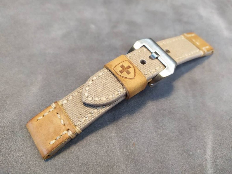 May include: A beige canvas watch strap with a brown leather trim and a silver buckle. The strap has a stitched design and a small shield with a cross logo.