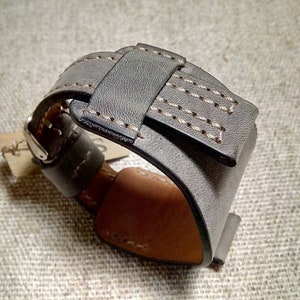 May include: A wide gray leather watch strap featuring a thick cuff design with visible light-colored stitching along the edges. The strap includes a metal buckle and a leather keeper loop, with a tan-colored interior lining and a small paper tag attached.