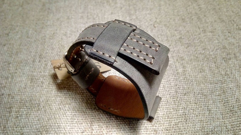 May include: A wide gray leather watch strap featuring a thick cuff design with visible light-colored stitching along the edges. The strap includes a metal buckle and a leather keeper loop, with a tan-colored interior lining and a small paper tag attached.