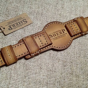 May include: A tan leather watch strap with a silver-coloured buckle. The strap has detailed stitching and the text "Leatherworks SHERP IGOR SHARAPA" is embossed on the central section. A small tag with the SHERP logo is attached.