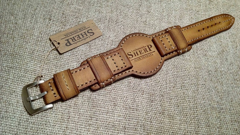 May include: A tan leather watch strap with a silver-coloured buckle. The strap has detailed stitching and the text "Leatherworks SHERP IGOR SHARAPA" is embossed on the central section. A small tag with the SHERP logo is attached.