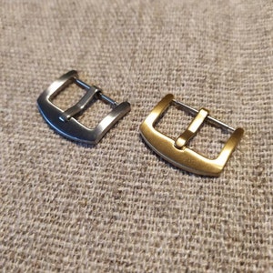 May include: Two watch buckles, one silver and one gold, rest on a textured, neutral surface. The buckles are rectangular with a single bar for the watch strap. The gold buckle has a warmer tone compared to the silver one. The background is a woven fabric.