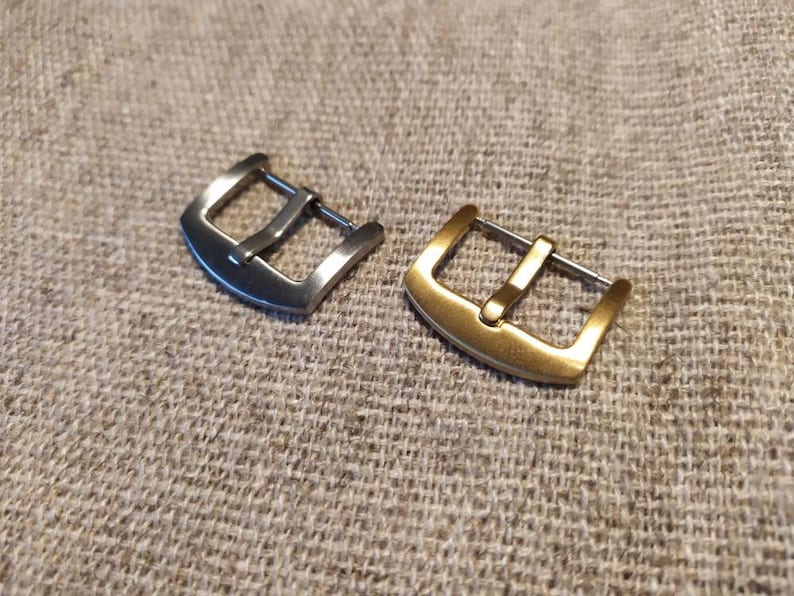 May include: Two watch buckles, one silver and one gold, rest on a textured, neutral surface. The buckles are rectangular with a single bar for the watch strap. The gold buckle has a warmer tone compared to the silver one. The background is a woven fabric.