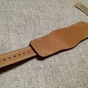 May include: A tan leather watch strap featuring a wide cuff design with visible stitching and a series of adjustment holes. A small rectangular tag attached to the end of the strap displays the text SHERP in black capital letters.