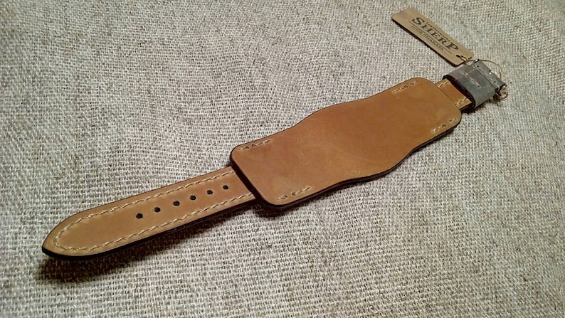 May include: A tan leather watch strap featuring a wide cuff design with visible stitching and a series of adjustment holes. A small rectangular tag attached to the end of the strap displays the text SHERP in black capital letters.