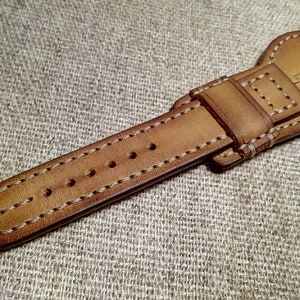 May include: A close-up shot of a tan leather watch strap with white stitching. The strap features a buckle and adjustment holes. The brand name "SHERE" is visible. The background is a textured, neutral-toned fabric.